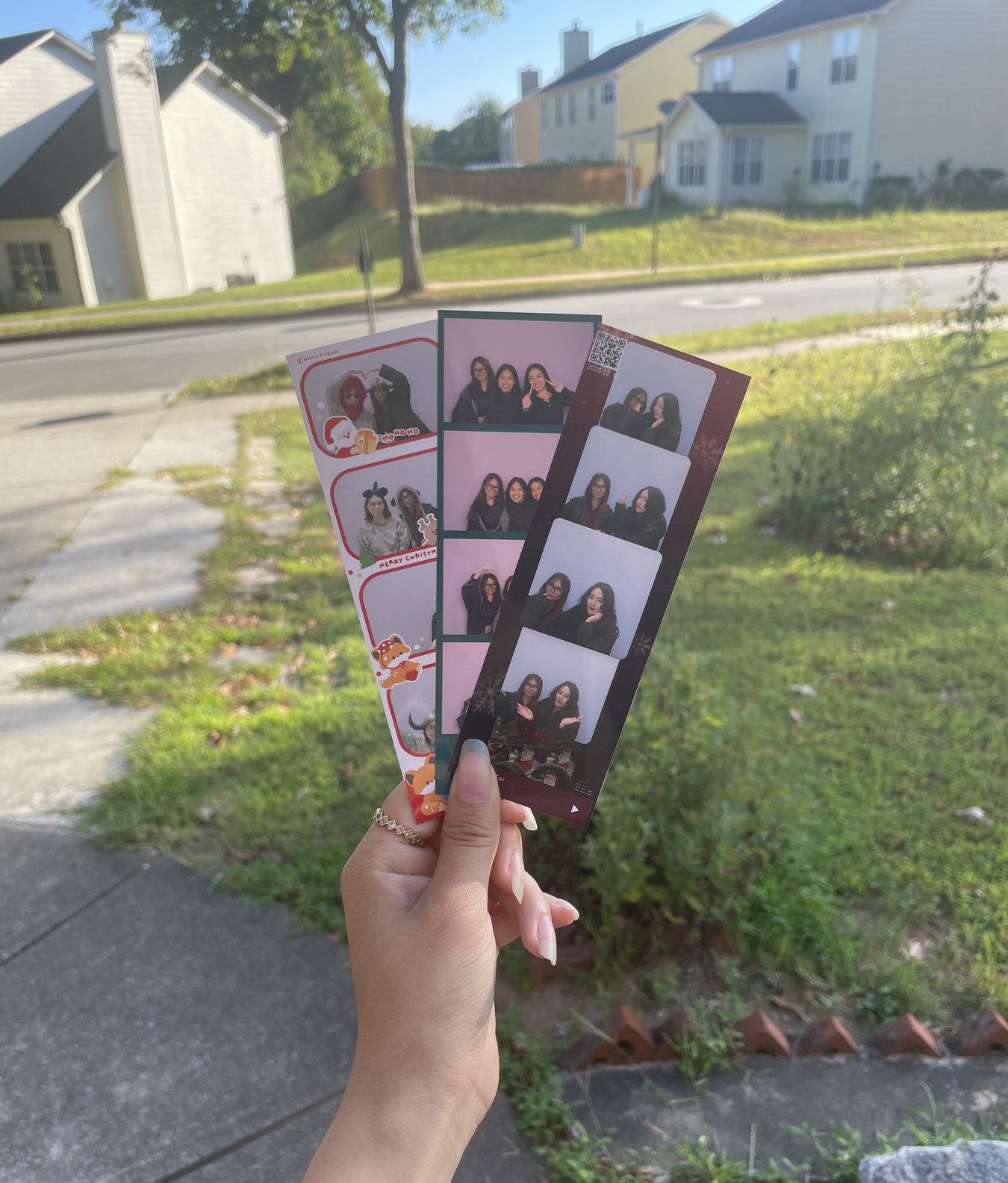 A hand holding up three photo-booth strips of friends outdoors on a sunny day